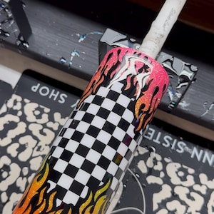 May include: A stainless steel tumbler with a vibrant design featuring a black and white checkered pattern and flames in shades of orange, yellow, and pink. The tumbler has a glittery finish.