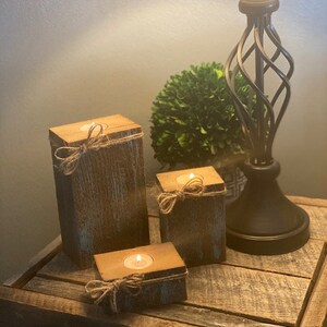 Puede incluir: Tres portavelas de madera r&uacute;stica con un acabado desgastado. Cada portavelas tiene una peque&ntilde;a vela de t&eacute; dentro y est&aacute; decorado con un trozo de cuerda atado alrededor de la parte superior.