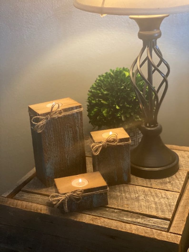 Puede incluir: Tres portavelas de madera r&uacute;stica con un acabado desgastado. Cada portavelas tiene una peque&ntilde;a vela de t&eacute; dentro y est&aacute; decorado con un trozo de cuerda atado alrededor de la parte superior.