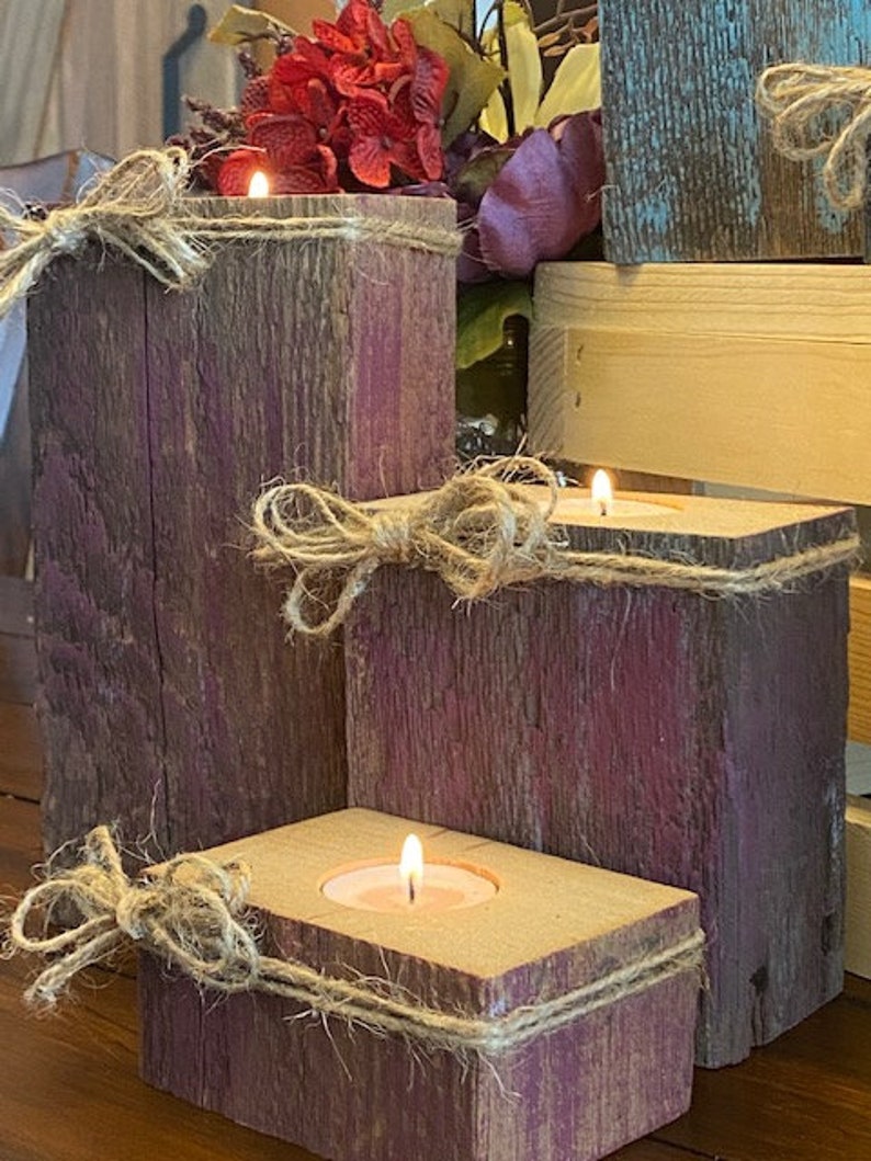 Puede incluir: Tres portavelas de madera r&uacute;stica con una mancha p&uacute;rpura y detalles de cuerda. Cada portavelas tiene una peque&ntilde;a vela de t&eacute;.