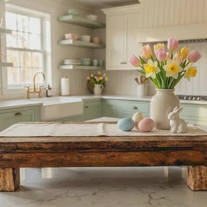 May include: A distressed wooden serving tray with a white linen runner, displaying a vase of tulips and daffodils, Easter eggs, and a white ceramic bunny. The tray is supported by four wooden blocks. The kitchen has a farmhouse style.