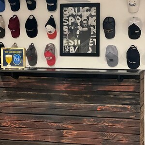 May include: A rustic wooden bar counter with a black countertop. The bar is made of reclaimed wood with a dark stain and has a metal bar rail. The bar is decorated with a collection of baseball caps hanging on the wall behind it. The wall is white and has a framed poster with the text "Bruce Springsteen" on it. There is a small sign on the counter that says "Think what happened to Piels".
