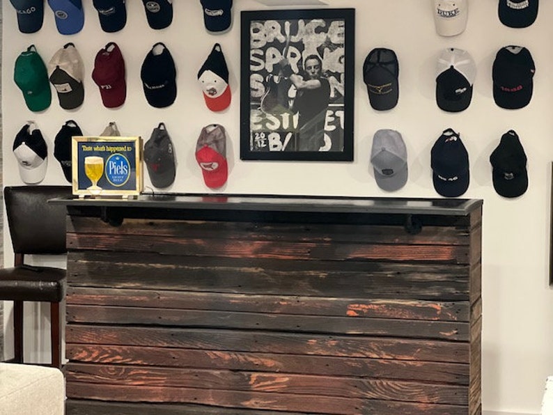 May include: A rustic wooden bar counter with a black countertop. The bar is made of reclaimed wood with a dark stain and has a metal bar rail. The bar is decorated with a collection of baseball caps hanging on the wall behind it. The wall is white and has a framed poster with the text "Bruce Springsteen" on it. There is a small sign on the counter that says "Think what happened to Piels".