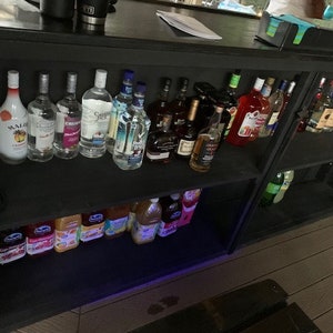 May include: A black wooden bar with two shelves, stocked with various bottles of liquor, including rum, vodka, and tequila. The bar also has a shelf with juice boxes, likely for making cocktails.