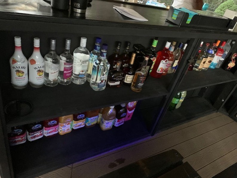 May include: A black wooden bar with two shelves, stocked with various bottles of liquor, including rum, vodka, and tequila. The bar also has a shelf with juice boxes, likely for making cocktails.