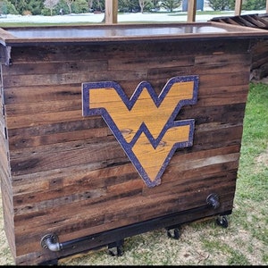 May include: A wooden bar cart with a dark brown finish and a West Virginia University logo. The cart has a black metal frame and wheels.
