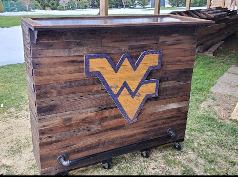 May include: A wooden bar cart with a dark brown finish and a West Virginia University logo. The cart has a black metal frame and wheels.