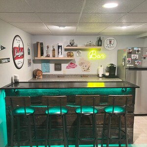 May include: A home bar with a wooden countertop and four bar stools. The countertop is lit with green LED lights. The bar is decorated with various signs and a neon sign that says "Bar".