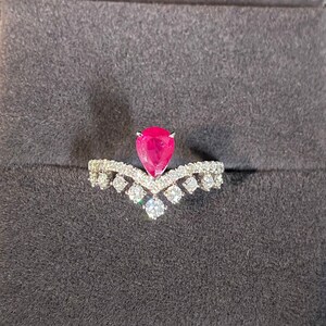 Könnte beinhalten: Ein Rubin- und Diamantring, präsentiert in einer grauen Schmuckschatulle. Der Ring hat einen birnenförmigen, leuchtend roten Rubin als Mittelpunkt, mit einem zarten Band, das mit kleinen Diamanten besetzt ist. Der Ring liegt auf einem dunkelgrauen Samtinnenraum.