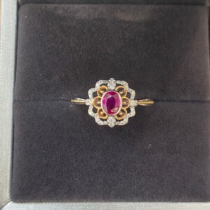 May include: A gold ring with a ruby center stone, surrounded by a floral design with white gemstones. The ring is displayed in a gray jewelry box with a dark gray interior.