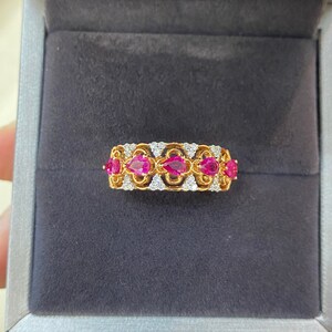 May include: A gold ring with a series of five ruby-colored gemstones in a teardrop shape, set in a gold band. The band is also adorned with small, clear gemstones. The ring is displayed in a gray jewelry box.