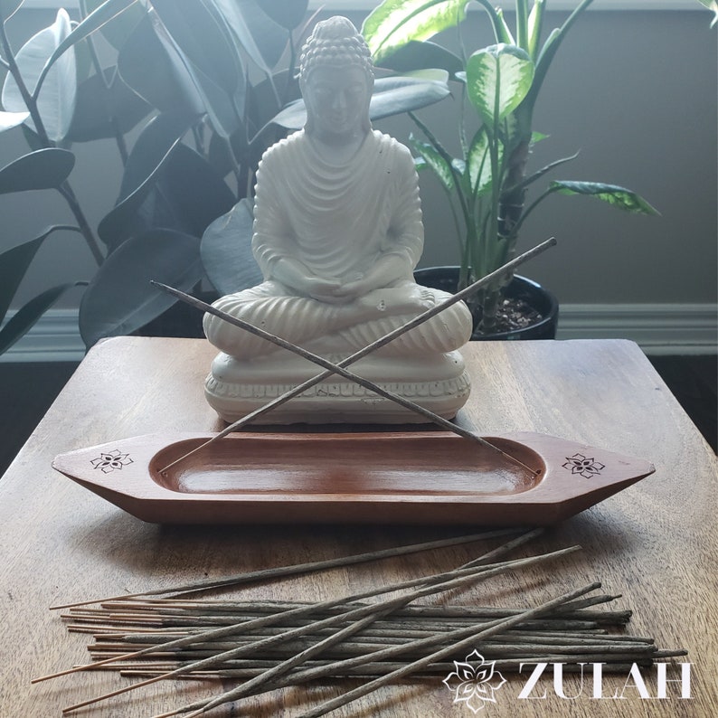 May include: A wooden incense holder shaped like a boat with a white ceramic Buddha statue sitting on a wooden surface. The incense holder has a carved floral design and is filled with gray incense sticks.