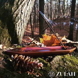 May include: A wooden incense burner with two incense sticks burning, creating smoke trails. The burner is resting on a mossy log in a forest setting with fallen leaves and a pine cone in the foreground. The text "ZULAH" is visible in the lower right corner.
