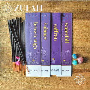 Peut inclure: Quatre boîtes rectangulaires violettes de bâtonnets d'encens de la marque Zulah étiquetées brown sugar, balance, saffron et waterfall. Chaque boîte contient 25 bâtonnets. Un faisceau de bâtonnets noirs à bouts orange et trois petites pierres polies.