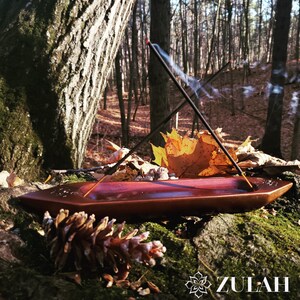 May include: A wooden incense burner with two incense sticks burning, creating smoke trails. The burner is resting on a mossy log in a forest setting with fallen leaves and a pine cone in the foreground. The text "ZULAH" is visible in the lower right corner.
