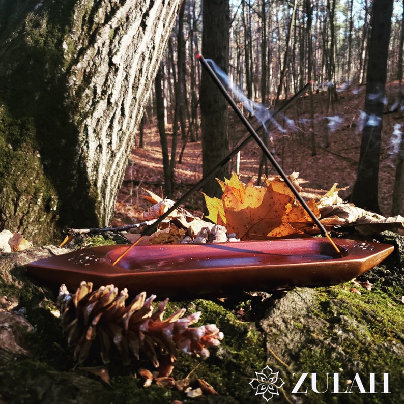 May include: A wooden incense burner with two incense sticks burning, creating smoke trails. The burner is resting on a mossy log in a forest setting with fallen leaves and a pine cone in the foreground. The text "ZULAH" is visible in the lower right corner.