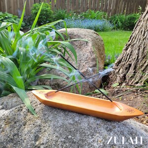 May include: A wooden incense burner shaped like a boat with two incense sticks burning inside. The burner is sitting on a large rock with green grass and a tree in the background.