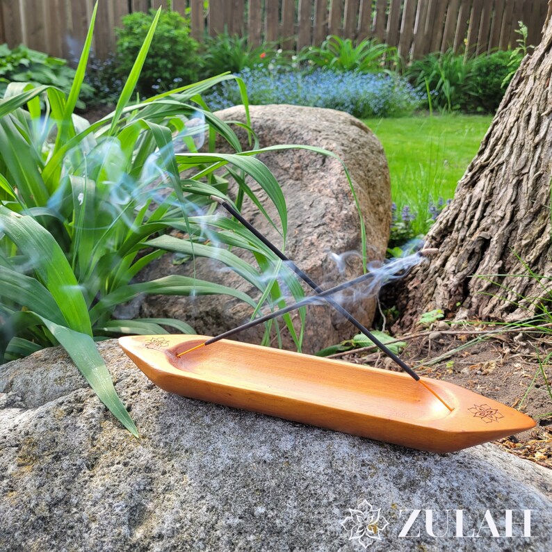May include: A wooden incense burner shaped like a boat with two incense sticks burning inside. The burner is sitting on a large rock with green grass and a tree in the background.