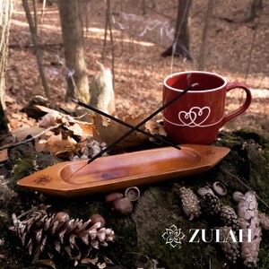 May include: A wooden incense burner with two incense sticks burning, a red mug with a white heart design, and a pine cone on a mossy surface.