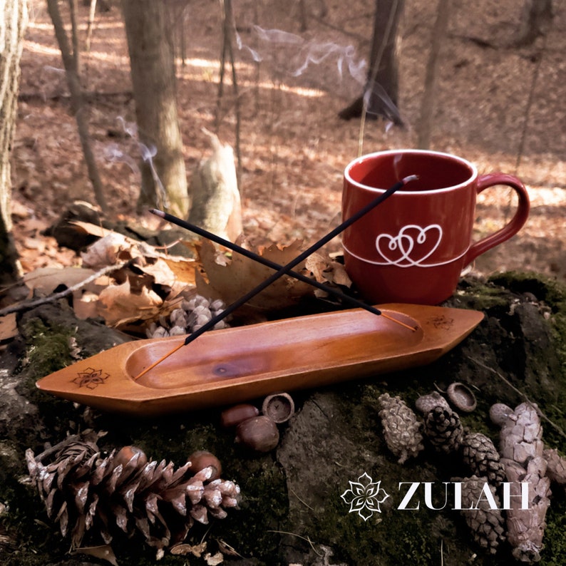 May include: A wooden incense burner with two incense sticks burning, a red mug with a white heart design, and a pine cone on a mossy surface.