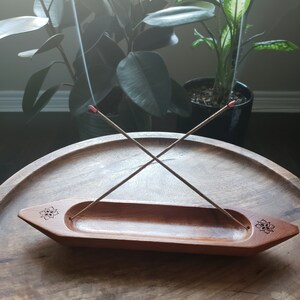 May include: A wooden incense holder shaped like a boat with two incense sticks burning. The holder has a carved floral design on each end.
