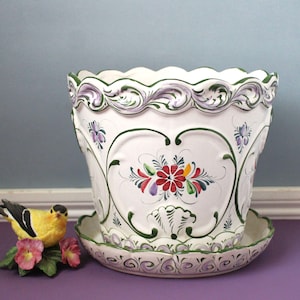 May include: A white ceramic planter with a matching saucer, featuring a scalloped rim and hand-painted floral motifs in red, green, and purple. The design includes embossed heart-shaped outlines and intricate scrollwork patterns.