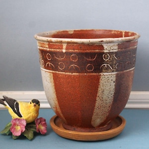 May include: A handmade ceramic plant pot with a rustic appearance. The pot has a warm colour scheme of brown, white, and terracotta, with a decorative band of etched patterns. It sits on a matching saucer, ideal for indoor plants.