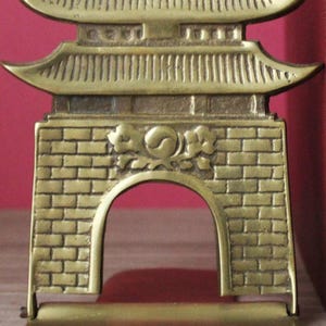 May include: A brass bookend featuring a detailed relief of a traditional gate structure with a curved roof and a central brick wall design. The center of the gate displays a circular emblem with a yin-yang symbol surrounded by floral motifs.