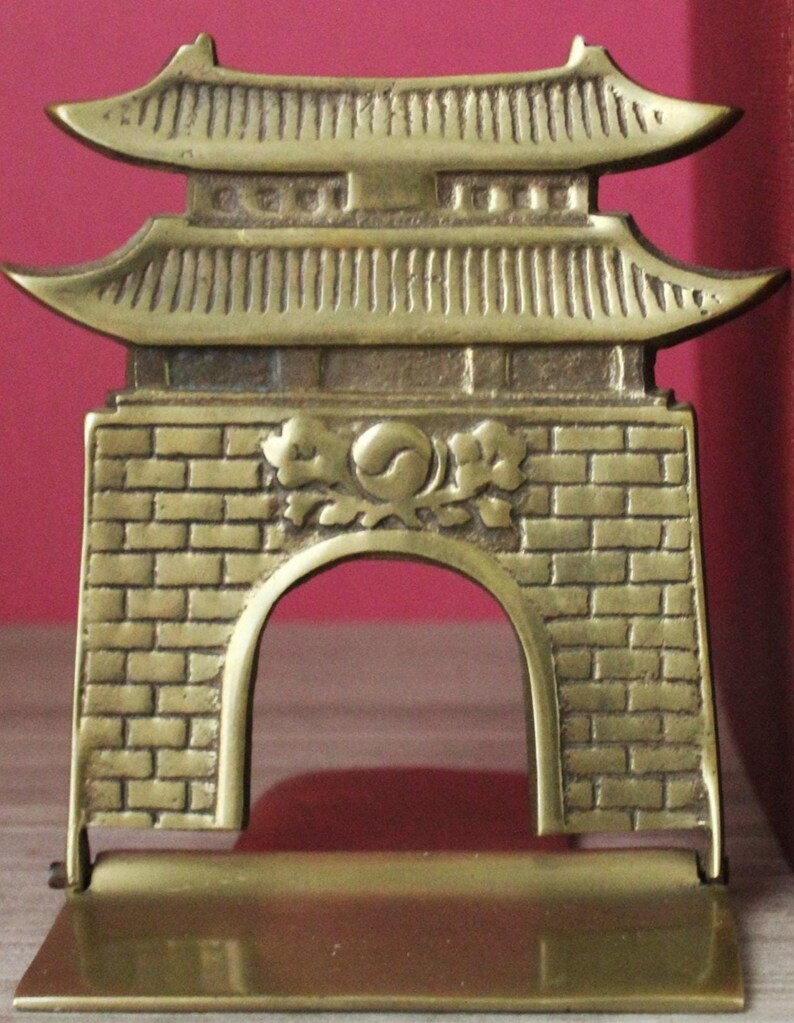 May include: A brass bookend featuring a detailed relief of a traditional gate structure with a curved roof and a central brick wall design. The center of the gate displays a circular emblem with a yin-yang symbol surrounded by floral motifs.
