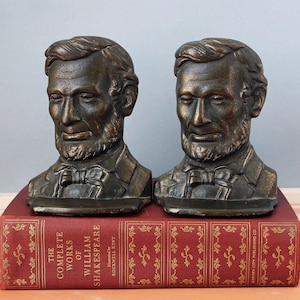 May include: A pair of bronze-toned metal bookends shaped as busts of Abraham Lincoln, each featuring detailed facial features, a beard, and a bow tie. The bookends are resting on a red hardcover book titled The Complete Works of William Shakespeare.