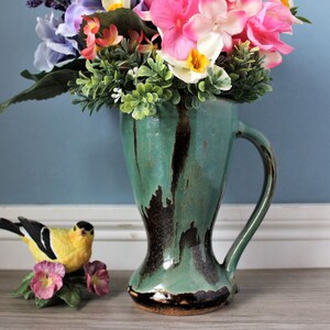 May include: A teal ceramic vase with a handle, filled with colorful artificial flowers. The vase has dark brown accents and a textured finish. A yellow bird figurine with pink flowers sits nearby.
