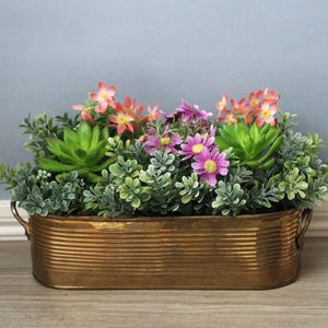 May include: An oval, ribbed, metallic bronze-colored planter with small side handles, filled with an arrangement of artificial green succulents, small grey-green foliage, and clusters of pink and purple daisy-like flowers.