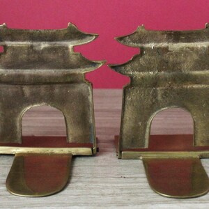 May include: A pair of brass bookends featuring a pagoda-style architectural design with a central arched opening. Each bookend has a flat, extended base plate and a vertical, multi-tiered roof structure with a weathered, metallic gold finish.
