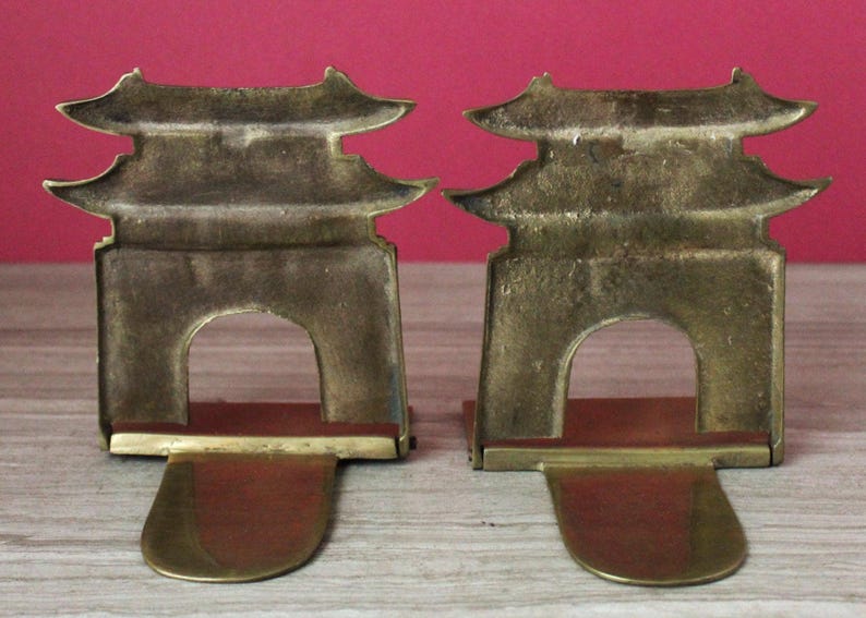 May include: A pair of brass bookends featuring a pagoda-style architectural design with a central arched opening. Each bookend has a flat, extended base plate and a vertical, multi-tiered roof structure with a weathered, metallic gold finish.