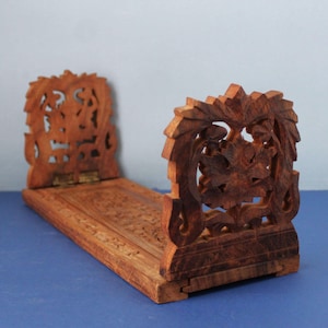 May include: A brown wooden book stand featuring intricate floral carvings on the two upright panels and the base. The stand includes metal hinges that allow the side panels to fold inward, showcasing detailed craftsmanship in the wood grain.