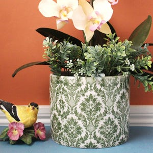 May include: A cylindrical ceramic planter featuring a green and white damask pattern. Inside the planter is an artificial orchid with white petals and green foliage. Beside the planter sits a small yellow and black bird figurine on pink flowers.