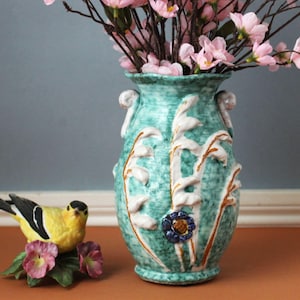 May include: A turquoise ceramic vase with a textured finish, featuring white raised floral relief patterns, gold accents, and two scroll handles. Beside it sits a yellow and black bird figurine perched on a base of green leaves and pink flowers.