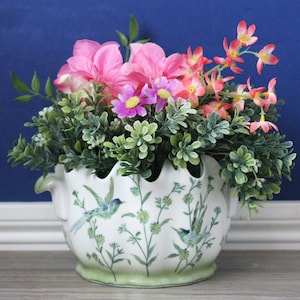 May include: A white ceramic planter with a scalloped rim and green base, decorated with painted green vines and blue birds. The planter is filled with an arrangement of artificial pink and coral flowers mixed with green foliage.