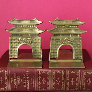 May include: A pair of brass bookends shaped like traditional East Asian gates with curved roofs and brick-patterned bases. Each gate features a central circular emblem. The bookends are resting on a red hardcover book titled The Complete Works of William Shakespeare.