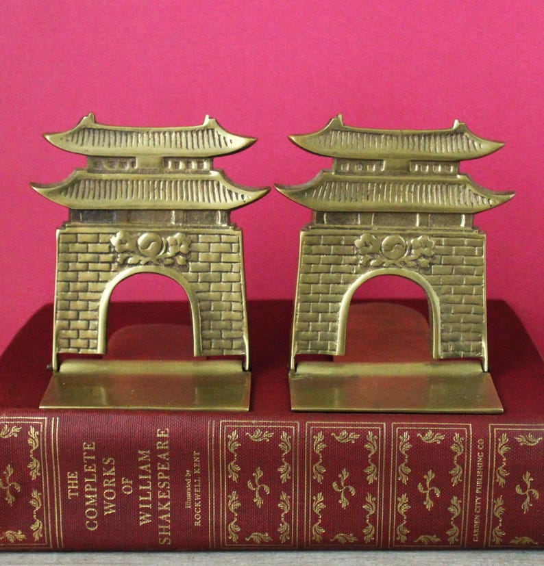 May include: A pair of brass bookends shaped like traditional East Asian gates with curved roofs and brick-patterned bases. Each gate features a central circular emblem. The bookends are resting on a red hardcover book titled The Complete Works of William Shakespeare.