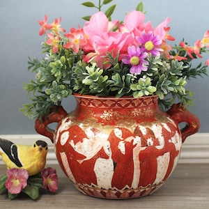 Può includere: Un vaso in ceramica rossa con due manici, decorato con figure dorate e bianche in abiti tradizionali, riempito con fiori artificiali rosa e viola. Una piccola statuetta di uccellino giallo e nero è posta accanto su una superficie in legno.