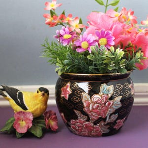 May include: A black ceramic planter featuring gold-outlined floral patterns and cloud motifs, filled with pink and yellow artificial flowers. Beside it sits a yellow and black bird figurine perched on a base of green leaves and pink flowers.