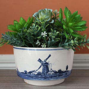 May include: A white ceramic planter with a blue windmill design, filled with green and blue artificial succulents and small white flowers. The planter has a rounded shape and a blue rim.
