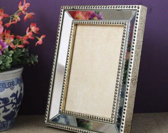 May include: A rectangular picture frame featuring a mirrored border with a beveled edge and a beaded inner trim. The outer frame has a textured, distressed silver finish. The center contains a plain, light beige insert for a photograph.