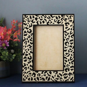 Può includere: Cornice fotografica rettangolare con un motivo leopardato nero e crema. La cornice ha un bordo nero e un interno color crema. Un piccolo vaso di fiori rosa e viola è visibile sullo sfondo.