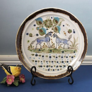 May include: A round ceramic plate with a dark brown rim featuring a hand-painted scene of two blue horses standing on a green grassy patch. Above the horses are stylized blue and green floral motifs, and below are rows of small blue dots.