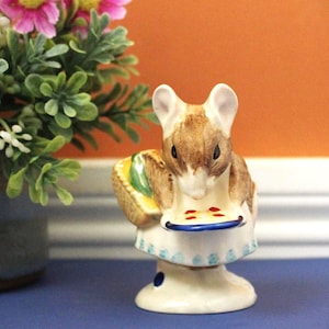 May include: A ceramic mouse figurine holding a pie, with a basket of food. The mouse is brown and white, wearing a white apron with blue accents. The figurine is on a pedestal base with a blue dot.