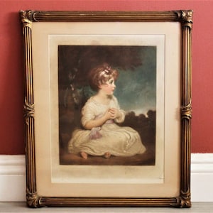 May include: A framed print of a young girl in a white dress, seated and looking to the side. The artwork is in a gold ornate frame with a cream-colored mat. The girl has a pink bow in her hair and a pink sash.