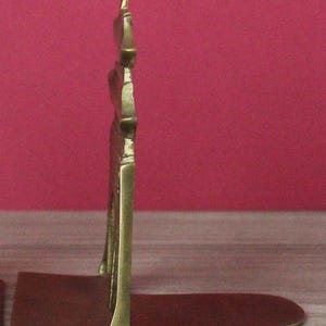 May include: A brass bookend featuring a vertical, tiered pagoda-style structure attached to a flat, horizontal base. The metal has a polished, golden-yellow finish with visible joints and a sturdy, L-shaped profile designed to support books upright.