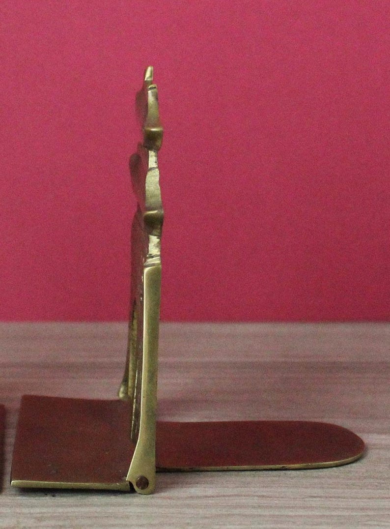 May include: A brass bookend featuring a vertical, tiered pagoda-style structure attached to a flat, horizontal base. The metal has a polished, golden-yellow finish with visible joints and a sturdy, L-shaped profile designed to support books upright.